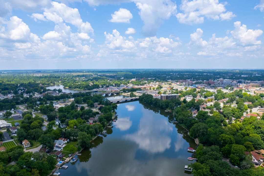 Aerial view of a tree-lined Illinois community surrounding a river under a bright, partly cloudy sky.
